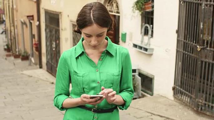 迷人的年轻黑发女人穿着浅色夏装在欧洲小镇的街道上。她正在看着手机并使用它。