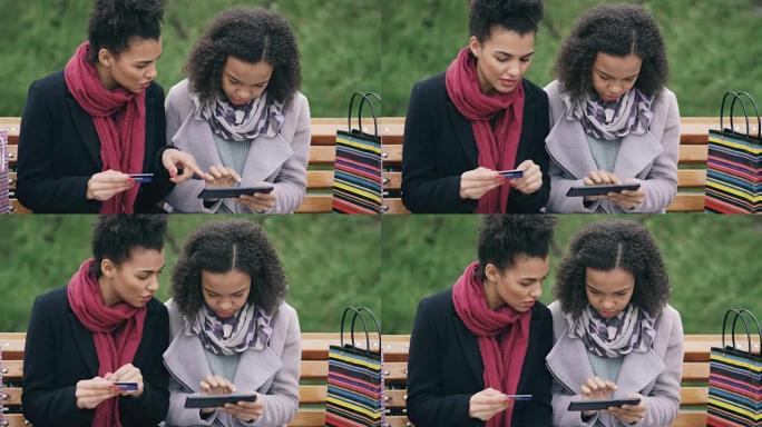 两个有吸引力的混血女人，纸袋坐在长凳上，使用平板电脑和信用卡进行在线购物