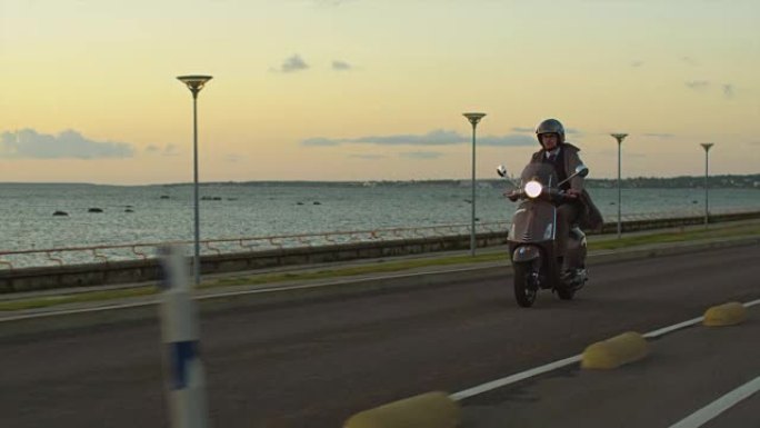 身穿优雅米色风衣的年轻人在日落时分在海边的道路上骑着复古踏板车。