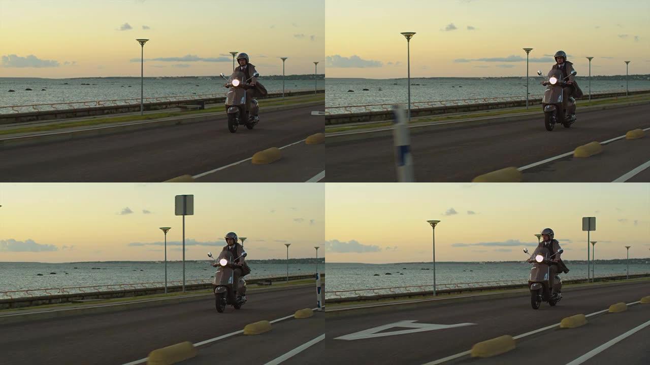 身穿优雅米色风衣的年轻人在日落时分在海边的道路上骑着复古踏板车。