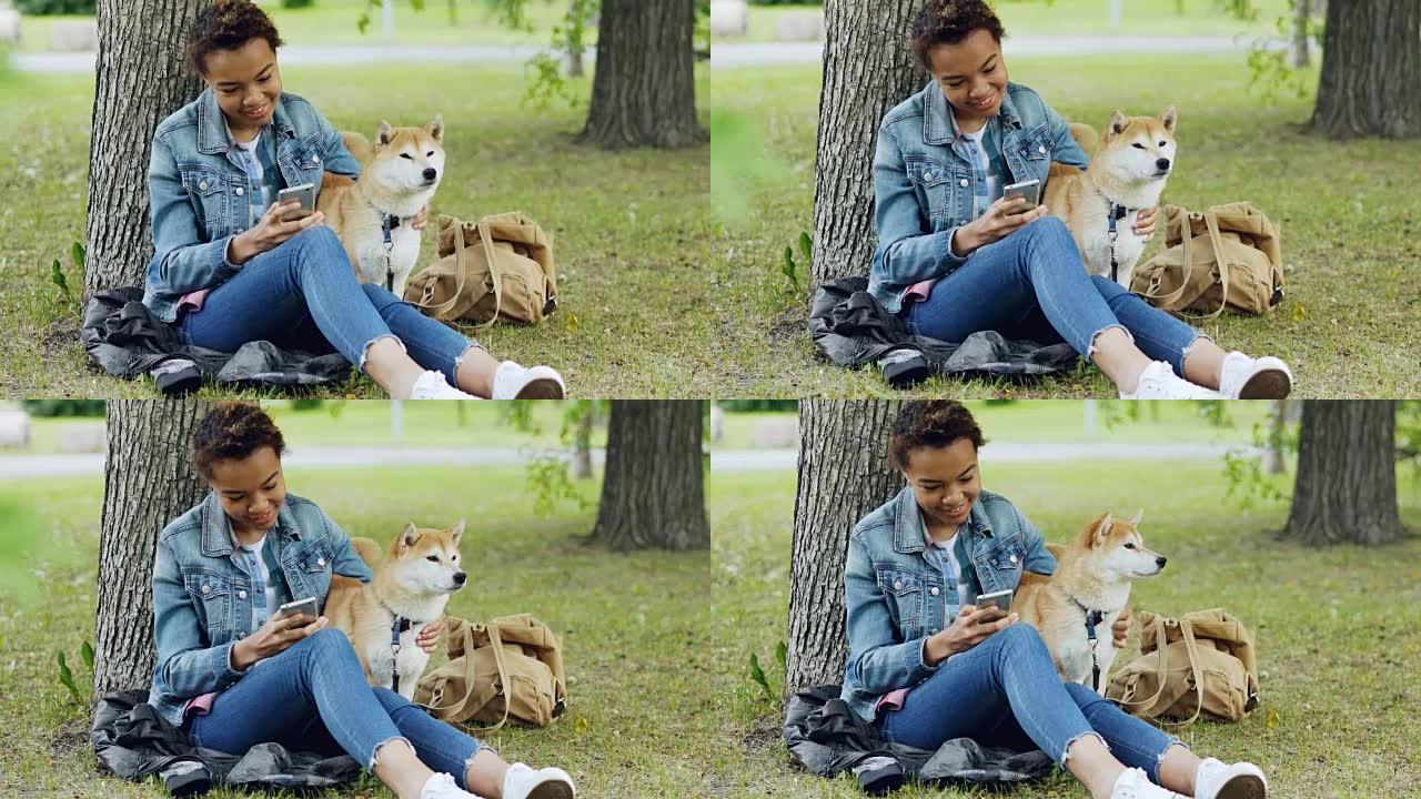 快乐的非洲裔美国妇女正在使用智能手机，在多风的夏日爱抚她可爱的宠物狗在城市公园休息。自然、动物和人的