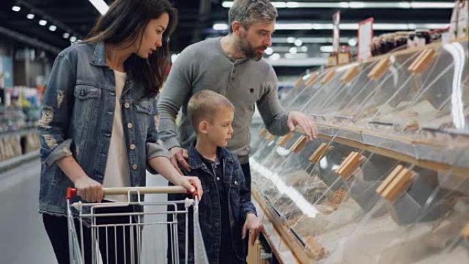 母亲，父亲和儿子正在超市里购物，指着架子上的塑料盒聊天。大型食品商店及其员工在背景中可见。