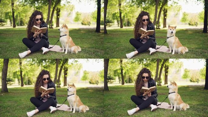 迷人的年轻女子正坐在公园的草地上看书，而她的良种狗正坐在主人附近环顾四周。休闲和动物概念。