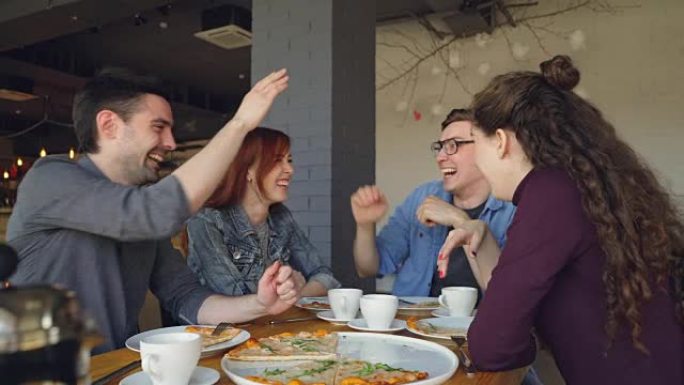 好看的学生在咖啡馆吃披萨时笑着做击掌和打手势。快乐的人，交流和美味的用餐概念。