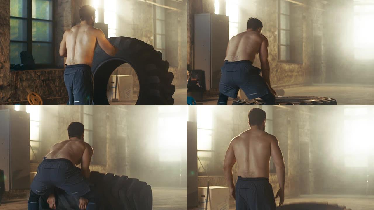 背部视图强壮的肌肉男子举起/翻转轮胎作为他的健身/交叉健身训练的一部分。他满身是汗，在一个废弃工厂改