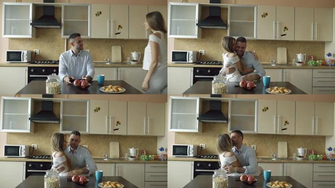 迷人的年轻夫妇清晨在厨房拥抱和亲吻，而男人吃早餐