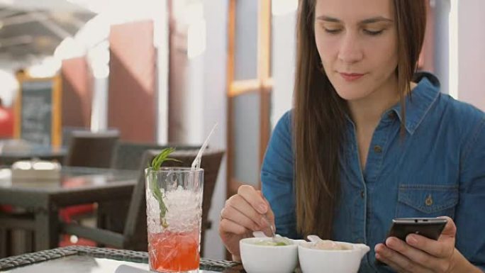 黑发女孩坐在咖啡馆外面吃冰淇淋时使用智能手机。全高清