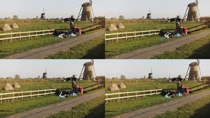 有两个孩子的妈妈坐在风车附近的草地上。夏日夕阳田园风光中宁静的家庭团聚。4K
