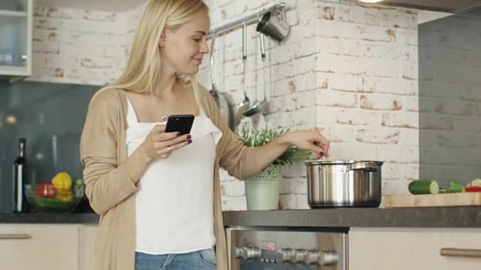 愉快的女人站在厨房前，用智能手机。她在锅里搅拌一些东西。