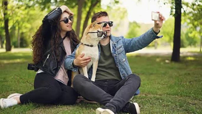 漂亮女人正在公园草地上休息时，用智能手机和她的男朋友和可爱的狗自拍。人类和动物都戴着墨镜。