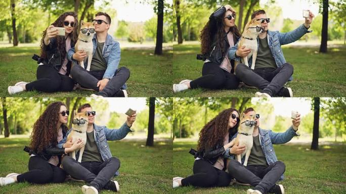 漂亮女人正在公园草地上休息时，用智能手机和她的男朋友和可爱的狗自拍。人类和动物都戴着墨镜。