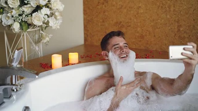 胡须上有泡沫的开朗男人正在现代水疗沙龙的热水浴缸中使用智能手机自拍。他在笑，打手势，摆姿势并看着相机