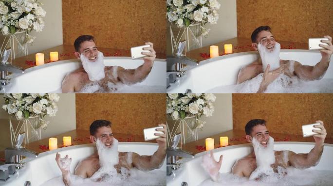 胡须上有泡沫的开朗男人正在现代水疗沙龙的热水浴缸中使用智能手机自拍。他在笑，打手势，摆姿势并看着相机