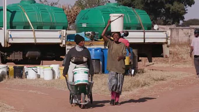 两名非洲妇女用塑料桶从一艘油轮上取水，然后走回自己的家