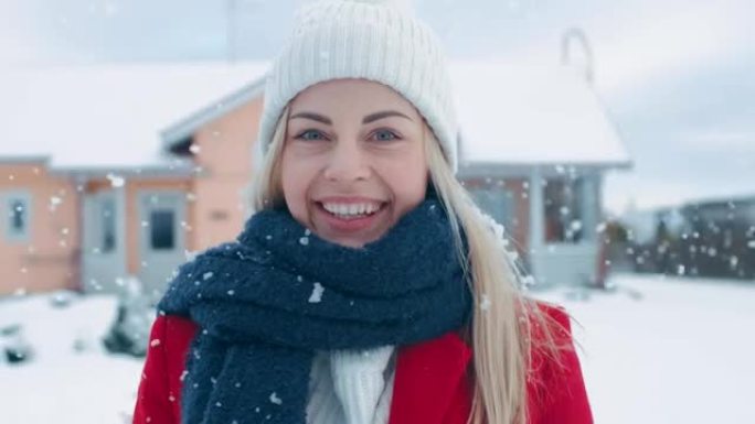 一位年轻的金发女子穿着红色外套，戴着围巾，站在飘落的雪中，对着镜头微笑。在后院的田园诗般的房子里，可