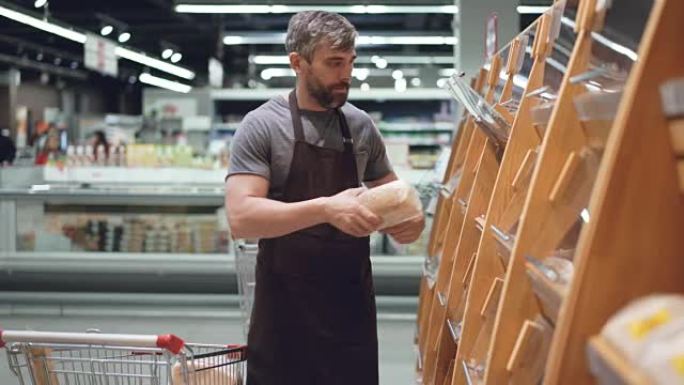 大型超市的男性员工正在从购物车中取出新鲜面包，并将其放在面包店部门的货架上。卖食物和人的概念。