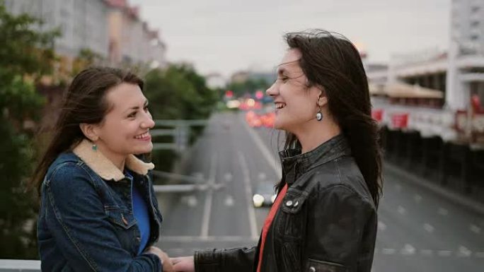 两个漂亮的女孩最好的朋友在城市大桥相遇，拥抱，亲吻，交谈，微笑，大笑。全高清