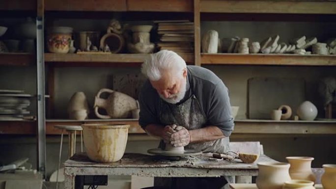 勤奋的白发男子正在陶工的轮子上使用粘土，塑造一片壤土。美丽的陶瓷器皿，手工制作的盆和架子上的花瓶是可
