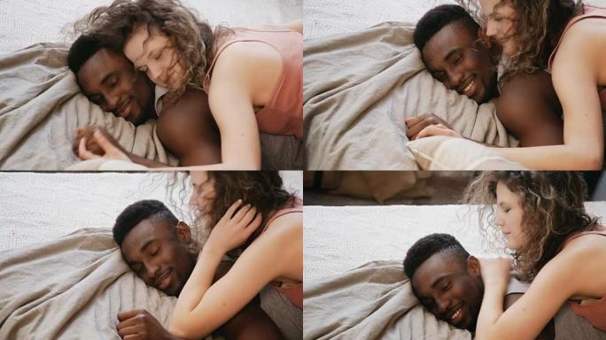 多民族夫妇浪漫的早晨。女人躺在男人身上，躺在床上拥抱。男女对话，牵手