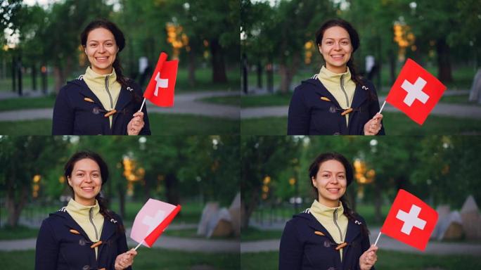 慢动作肖像快乐的瑞士女孩挥舞瑞士国旗，微笑着看着相机。背景是美丽的公园，有高高的树木和绿色的草坪。