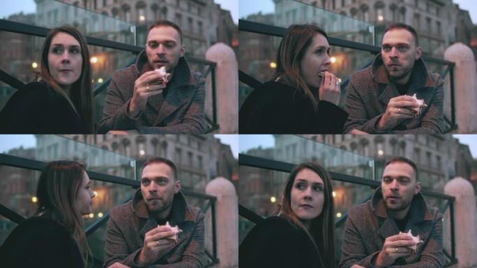 年轻的女人坐在外面，男人来了，坐在她旁边。迷人的夫妇吃三明治和聊天