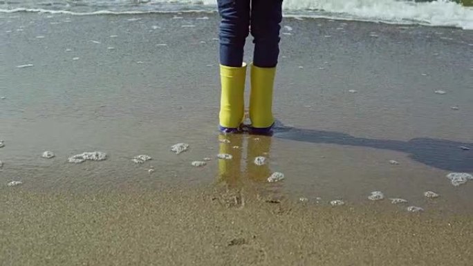 穿着亮黄色橡胶靴的小女孩脚的特写镜头。孩子站在沙滩上，脚印在沙滩上