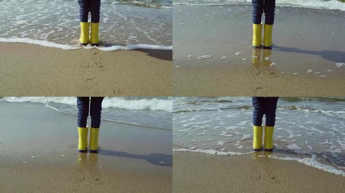 穿着亮黄色橡胶靴的小女孩脚的特写镜头。孩子站在沙滩上，脚印在沙滩上