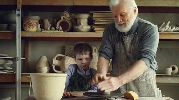 富有爱心的银发祖父正在小作坊教年轻可爱的孙子在掷轮上与黏土一起工作。陶器、家庭爱好和手工艺概念。
