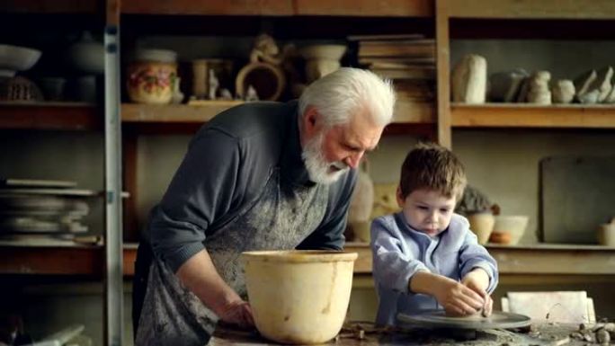 陶艺老师资深成人正在帮助年轻学生用抛掷轮上的一块粘土制成锅。可爱的男孩很笨拙，工匠正在帮助他。
