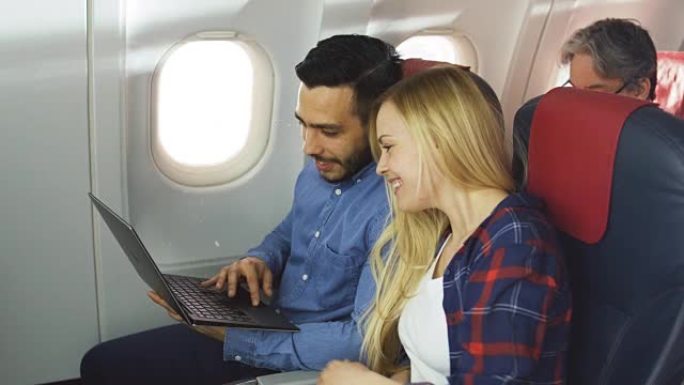 在商用飞机上，漂亮的年轻金发女郎和英俊的西班牙裔男性在笔记本电脑上看电影并大笑。阳光透过飞机的窗户照