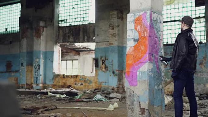 年轻的涂鸦艺术家正在用喷雾涂料在旧肮脏建筑的柱子上绘画。苛刻的家伙穿着蓝色牛仔裤，夹克和防护手套。
