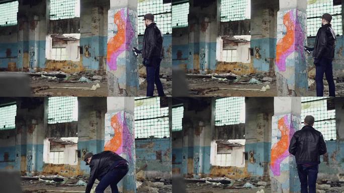 年轻的涂鸦艺术家正在用喷雾涂料在旧肮脏建筑的柱子上绘画。苛刻的家伙穿着蓝色牛仔裤，夹克和防护手套。