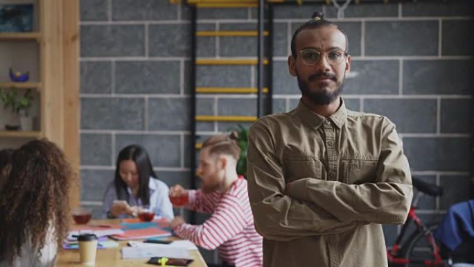 年轻的非洲裔美国商人的肖像，穿着衬衫和眼镜微笑着看着相机，而他的同事在室内现代办公室工作