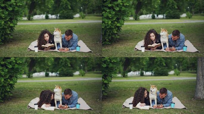 年轻的家庭在公园度过周末，女人看书，男人使用智能手机，可爱的狗在他们之间休息。这家伙正在给他妻子看手