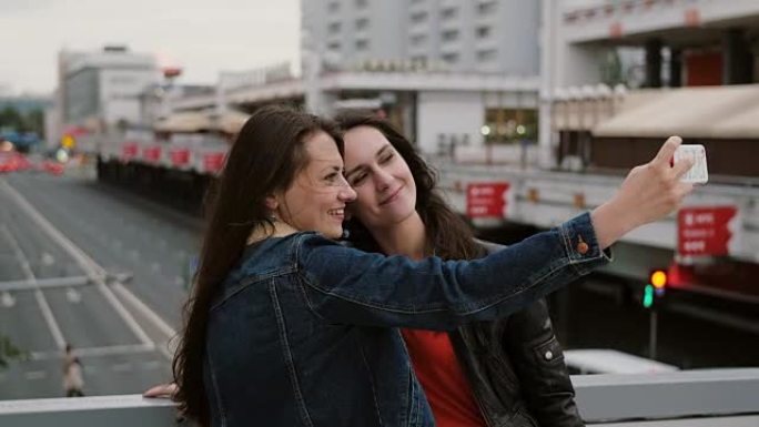 两个女孩最好的朋友自拍，站在城市的桥上，说话，微笑，大笑。全高清