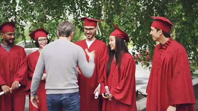 大胡子男人骄傲的老师祝贺毕业生在大学校园里与他们握手并拥抱他们，而学生们正在交谈并持有文凭。