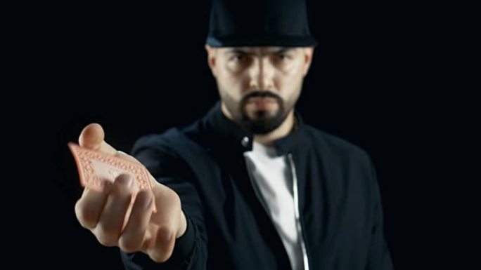 戴着帽子的专业街头魔术师表演手牌技巧，用手指旋转一张牌。背景是黑色的。