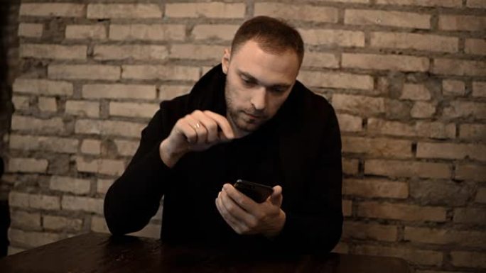 年轻的美男子坐在靠近砖墙的咖啡馆里，使用智能手机，独自上网