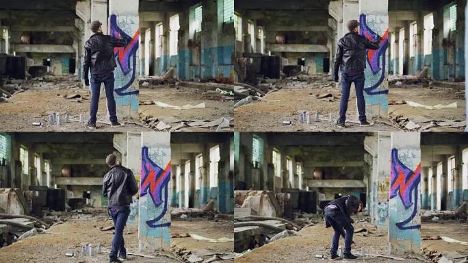 涂鸦画家在废弃建筑内用气溶胶涂料创造美丽形象的后视图。艺术家穿着蓝色牛仔裤，黑色皮夹克，帽子和手套。