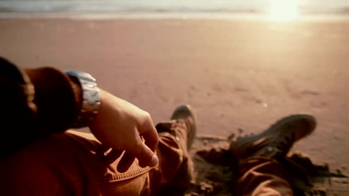 一个男人坐在沙滩上穿着靴子看着美丽的日落的第一视点视频