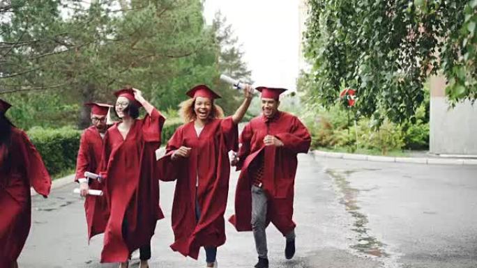 兴奋的即将毕业的学生穿着长袍和传统帽子在校园里带着文凭跑步，正在下雨。高等教育和幸福观。