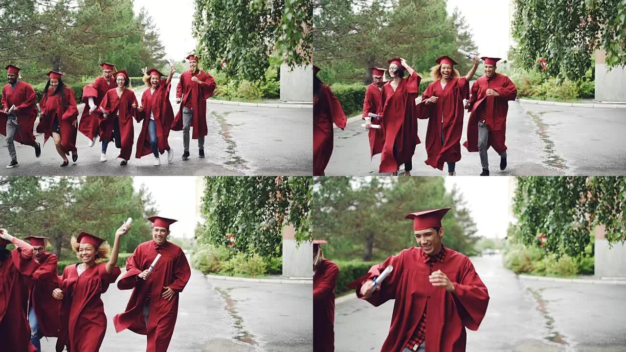 兴奋的即将毕业的学生穿着长袍和传统帽子在校园里带着文凭跑步，正在下雨。高等教育和幸福观。