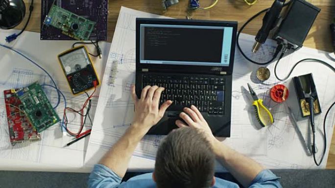 一位有天赋的it技术人员在笔记本电脑上编写代码的俯视图坐在办公桌前，他被各种技术组件所包围。阳光照在