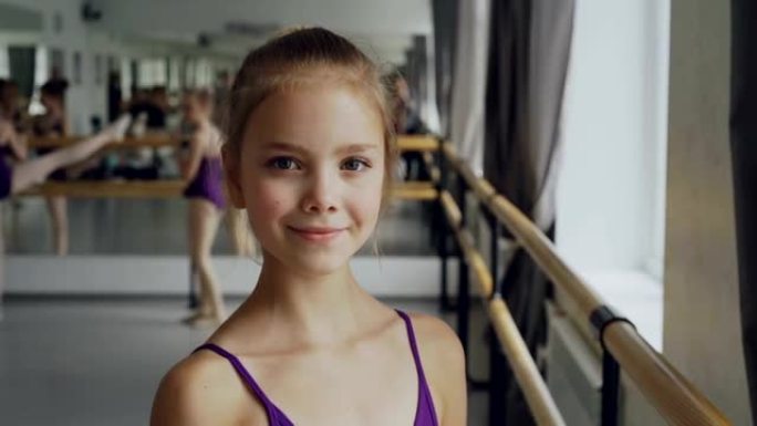 穿着紧身衣的美丽小女孩的特写肖像站在芭蕾舞课上，微笑着看着相机。其他学生正在做背景练习。