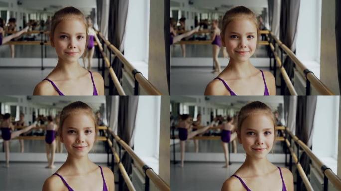 穿着紧身衣的美丽小女孩的特写肖像站在芭蕾舞课上，微笑着看着相机。其他学生正在做背景练习。