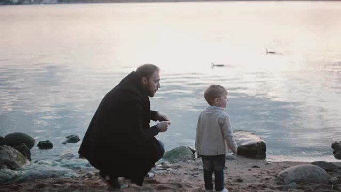 父亲和儿子在岸上交谈。男子蹲着，向男孩展示如何玩跳石。小男孩为喜悦而跳跃