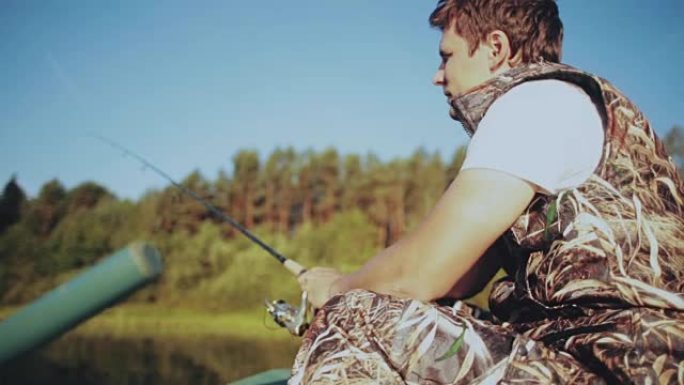 孤独的年轻人坐在橡皮船上，在湖边钓鱼。男性拿着钓鱼竿，扭转旋转卷轴