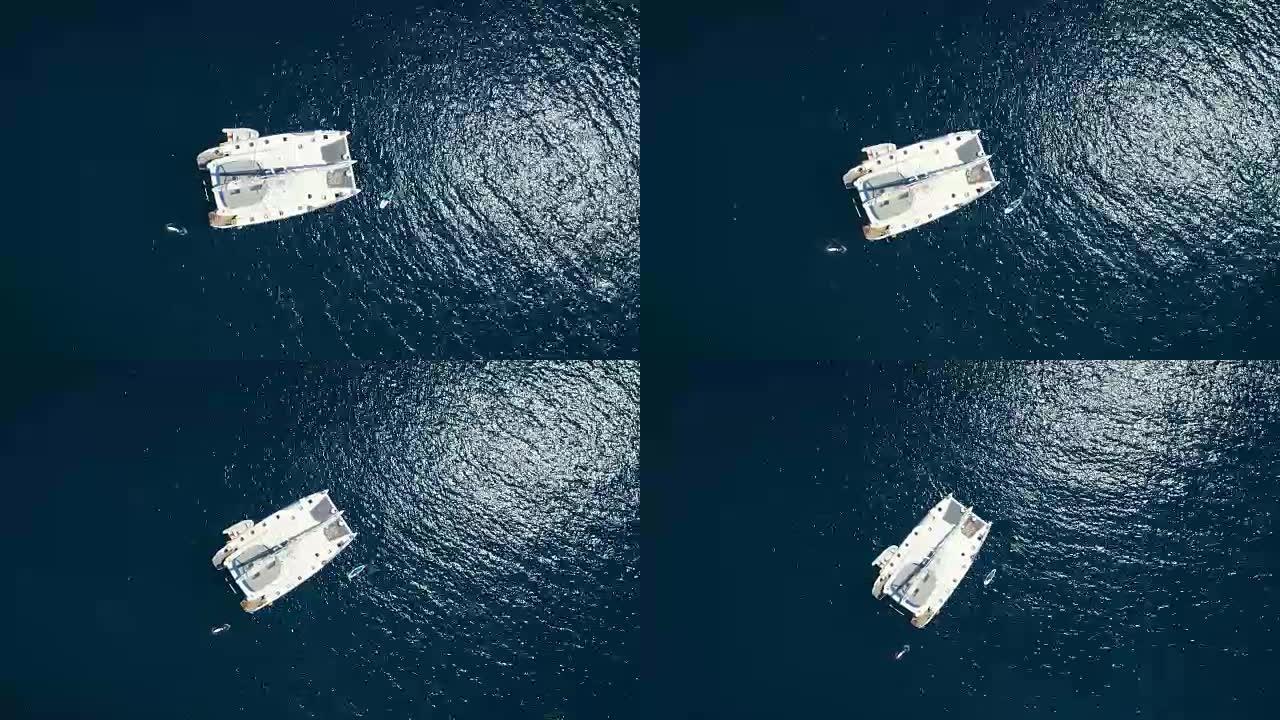 一艘大型白色帆船游艇的空中俯拍，停泊在海湾中，附近有站立式桨板。海洋是深蓝色的，美丽的。
