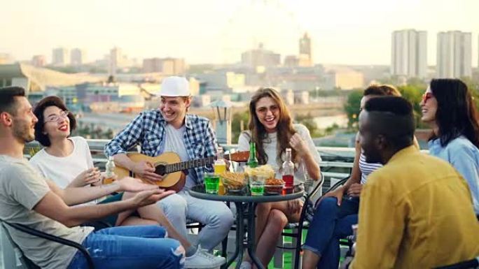 男吉他手正在弹吉他，而他的朋友们在屋顶派对上唱歌，大笑，喝啤酒和软饮料。现代城市在背景中可见。