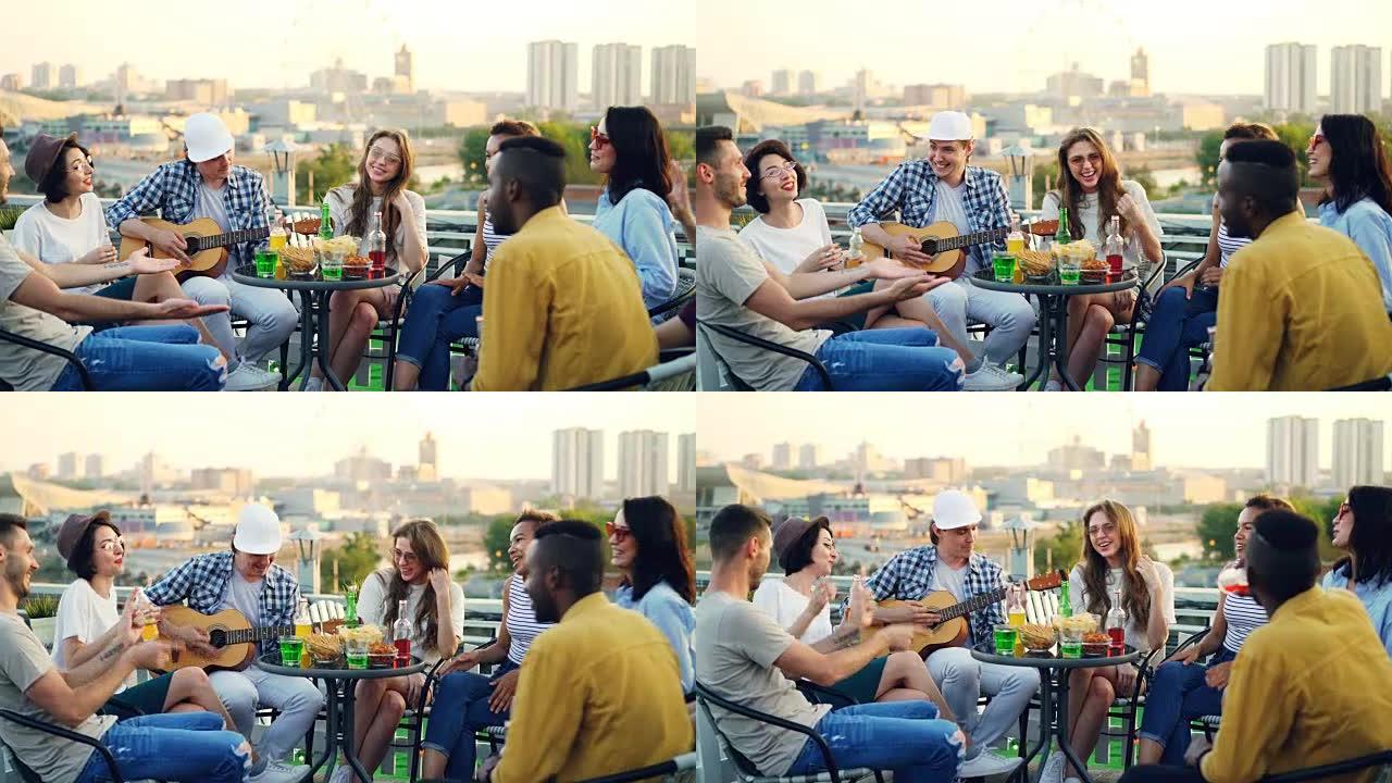 男吉他手正在弹吉他，而他的朋友们在屋顶派对上唱歌，大笑，喝啤酒和软饮料。现代城市在背景中可见。